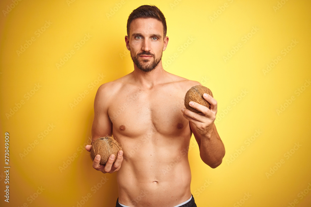 Young handsome shirtless man holding exotic tropical coconut over isolated yellow background with a confident expression on smart face thinking serious