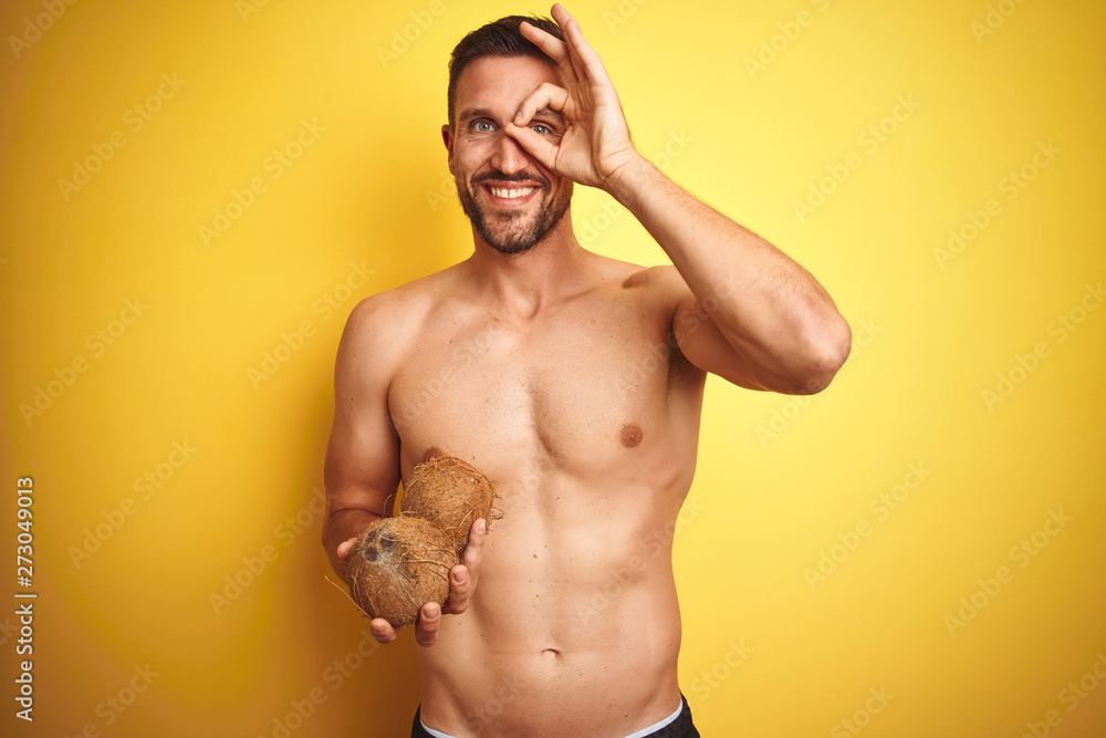 Young handsome shirtless man holding exotic tropical coconut over isolated yellow background with happy face smiling doing ok sign with hand on eye looking through fingers