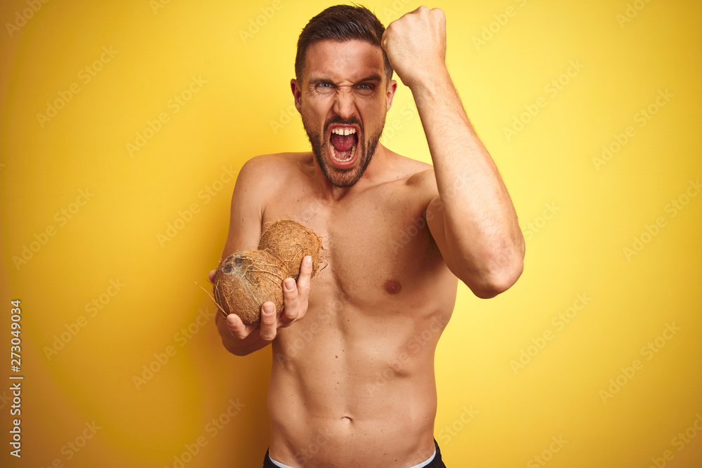 Fototapeta premium Young handsome shirtless man holding exotic tropical coconut over isolated yellow background annoyed and frustrated shouting with anger, crazy and yelling with raised hand, anger concept