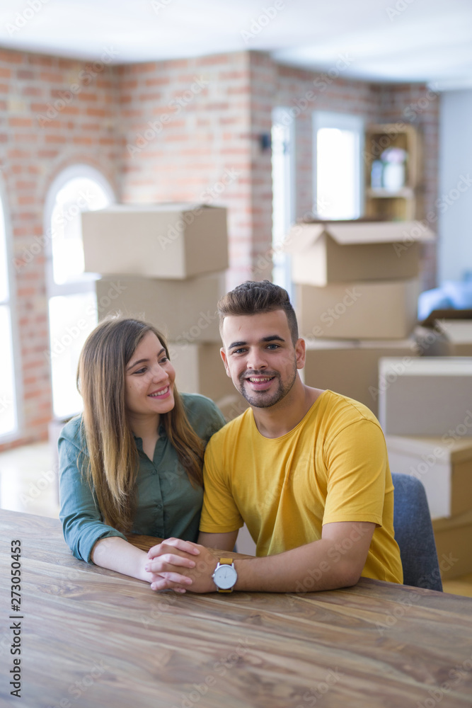 Young beautiful couple sitting on the table at home, hugging in love very happy for moving to new home with cardboard boxes behind them Young beautiful couple sitting on the table at home, hugging in love very happy for moving to new home with cardboard boxes behind them