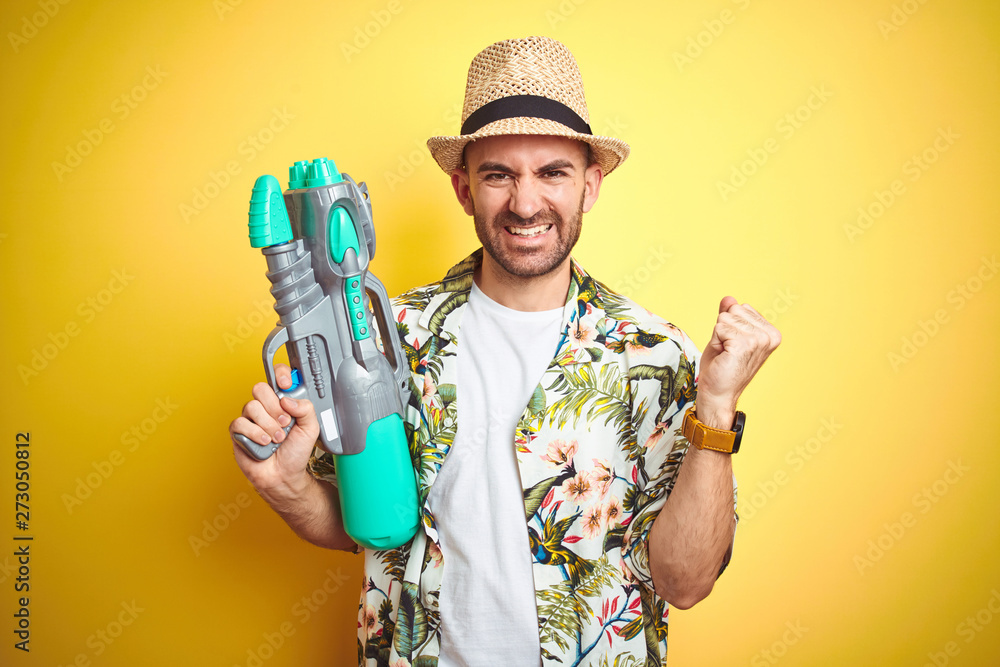 Young man wearing hawaiian flowers shirt holding water gun over yellow isolated background screaming proud and celebrating victory and success very excited, cheering emotion