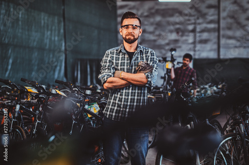 Pensive man in protective glasses and checkered shirt is posing at his own warehouse full of bicycles.