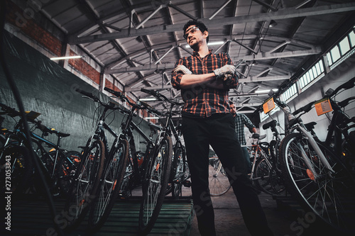 Attractive pensive mam in glasses is posing next to bicycles at his own warehouse.