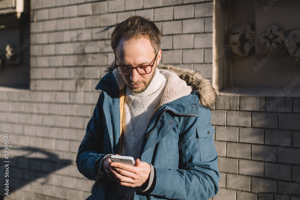 Pleasant male with beard, glasses and in blue jacket looking into mobile phone and drinking coffee. Social media addiction, dependency on a cell phone. Chatting, flirting and surfing in web on the go. Pleasant male with beard, glasses and in blue jacket looking into mobile phone and drinking coffee. Social media addiction, dependency on a cell phone. Chatting, flirting and surfing in web on the go.