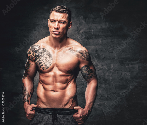Studio portrait of suntanned shirtless muscular male with tattoo on his chest over grey background.