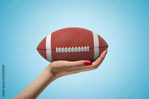 Side closeup of woman's hand facing up and holding big brown ball for American football on light blue gradient background.