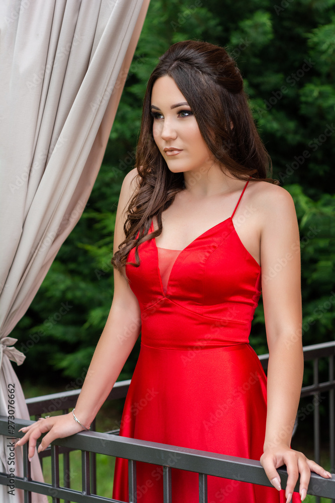 Attractive girl in red dress posing behind the fence on the balcony and curtains in the restaurant garden on a summer day. Fashion and beauty concept. Close up, selective focus Attractive girl in red dress posing behind the fence on the balcony and curtains in the restaurant garden on a summer day. Fashion and beauty concept. Close up, selective focus