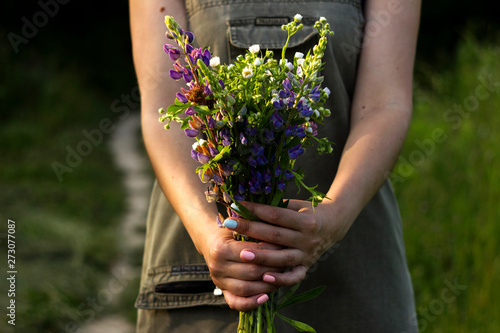 A young woman with a bouquet of wild flowers (chamomile, lupine, clover) in her hands is standing in a meadow, close-up. Summer, background, concept