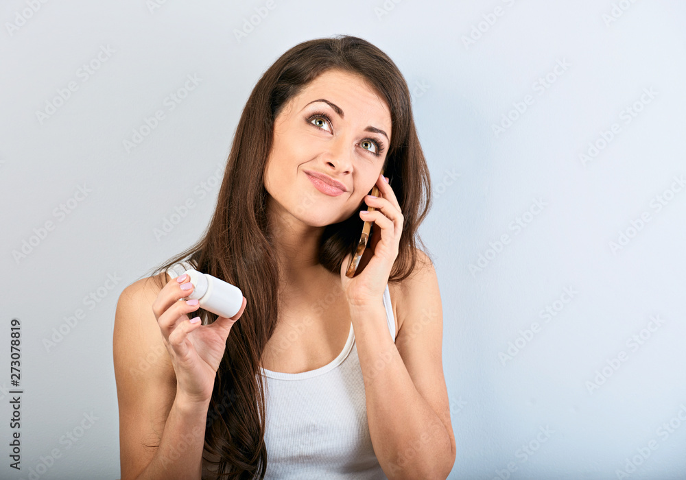 Fototapeta premium Worried confused young woman holding tablet bottle in the hand and calling on mobile phone to ask the doctor. She is looking up on blue background. Closeup