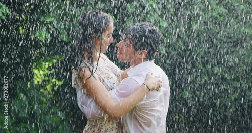 Slow motion of young carefree couple in love are dancing and kissing under the rain on a background of green trees.