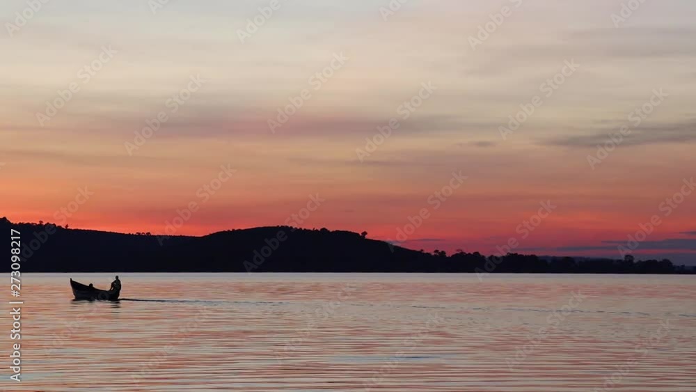 Silhouette of small fishing boat heading out to sea under orange sky after sunset at Lake Victoria. Boat slowly moves out of frame. Kalangala, Uganda.