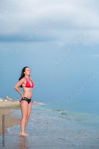 a slim brunette in a bikini stands on a sandy beach near the surf whit hands on her waist
