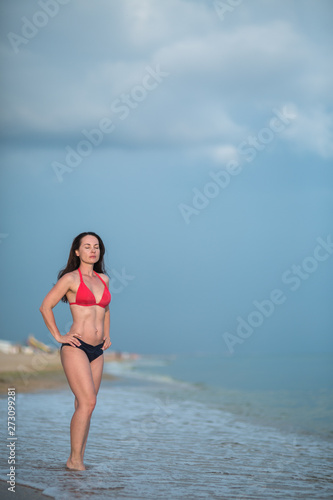 a slender brunette in a bikini stands on a sandy beach near the surf whit hands on her waist