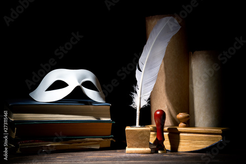 Drama or theater and literature concept. Old inkstand with feather near scrolls with mask on books against black background. Dramatic light