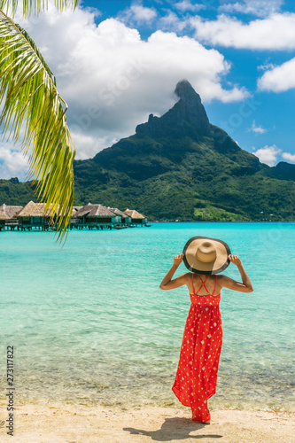Bora Bora Tahiti exotic tropical vacation destination tourist woman on ocean beach at view of Mt Otemanu in French Polynesia. Girl in red dress and hat relax on honeymoon getaway.