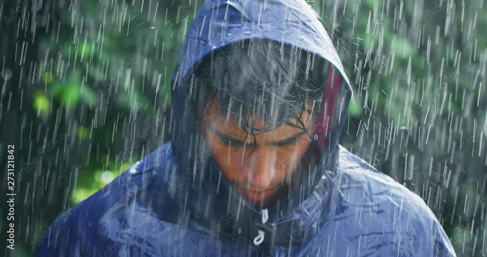 Slow motion of young carefree handsome young man wearing protection cape is feeling free under the rain on a background of green trees.