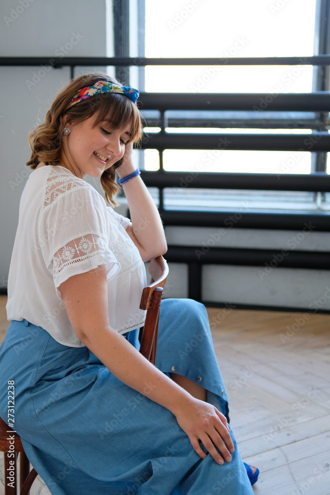 A young plus size woman sits on a chair in a spacious white room. Smile on his face, flirtatious look. Happiness and beauty in any weight. A girl in a long skirt, sandals with high heels A young plus size woman sits on a chair in a spacious white room. Smile on his face, flirtatious look. Happiness and beauty in any weight. A girl in a long skirt, sandals with high heels