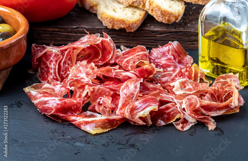 Appetizing slices Iberian ham in the foreground. Olive oil, bread, fresh tomato, olives. Black background. Rustic and homemade appearance.
