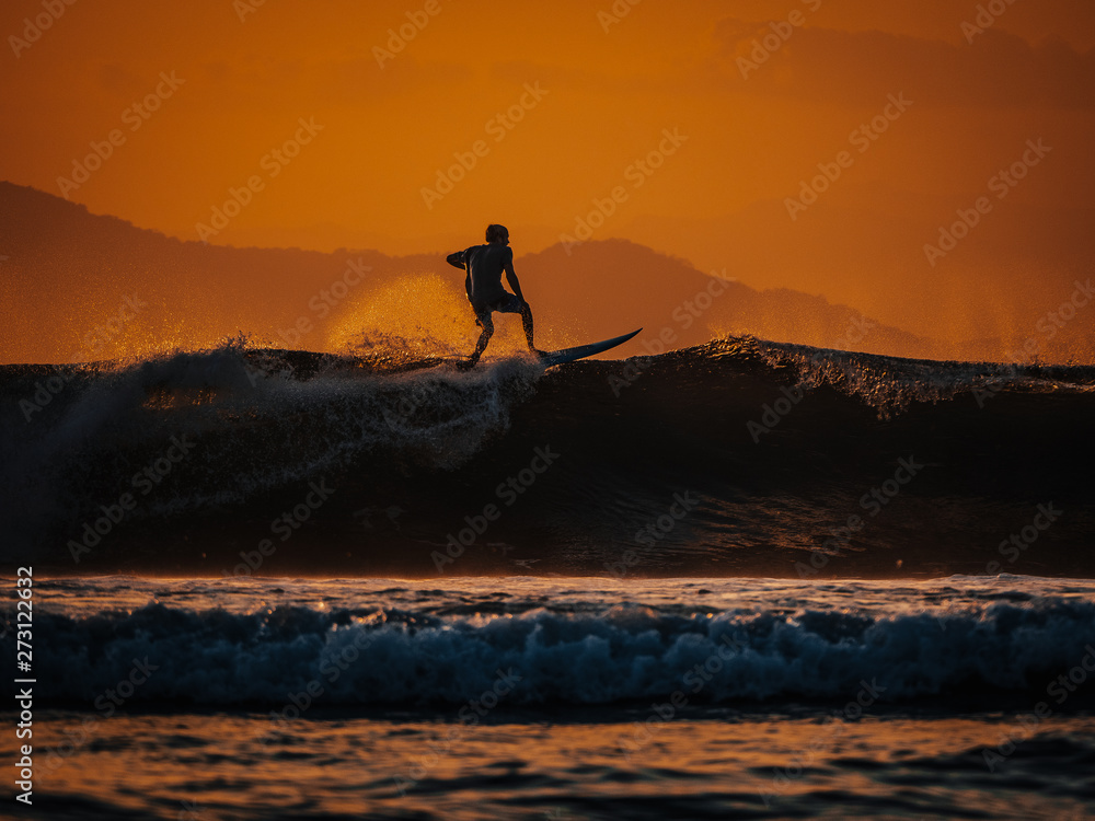 Silhouette of male surfer riding big ocean wave at orange sunset 
