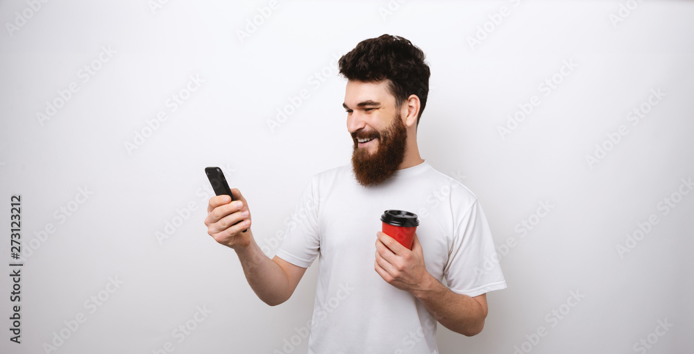 cheerful young bearded man surfing the internet on his phone and holding a red paper cup. cheerful young bearded man surfing the internet on his phone and holding a red paper cup.