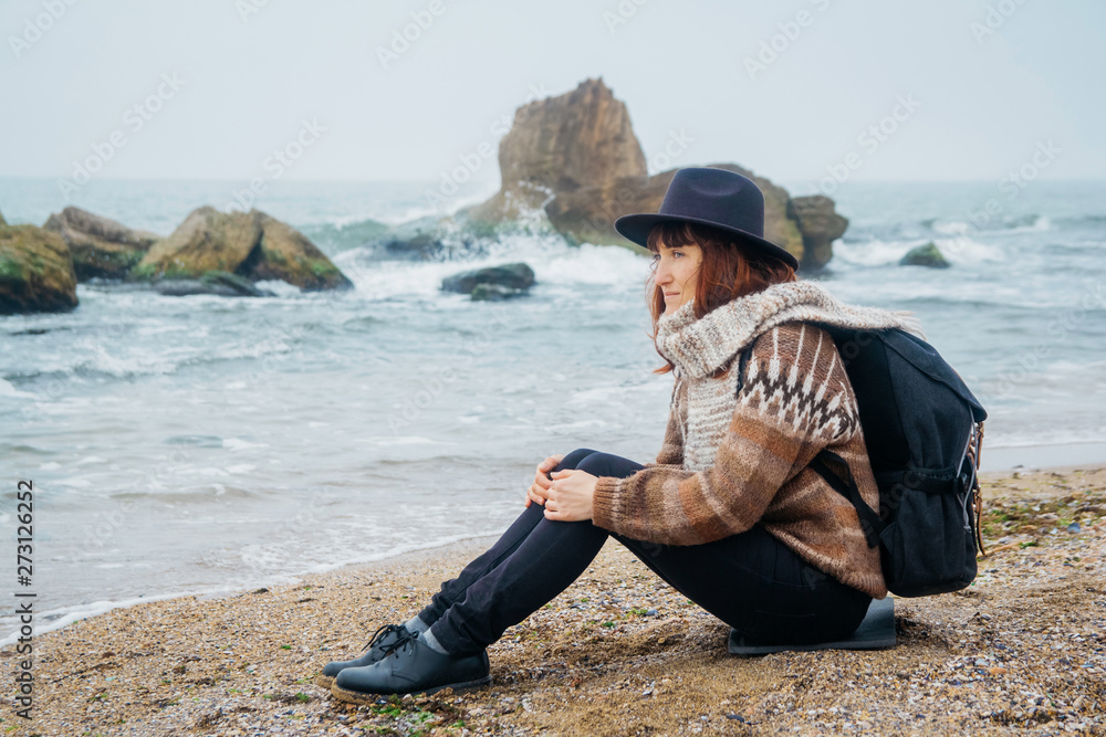 Beautiful red-haired woman in a hat and scarf with a backpack sits on the coast against the background of the rocks against the beautiful sea. Tourism, rest, lifestyle.