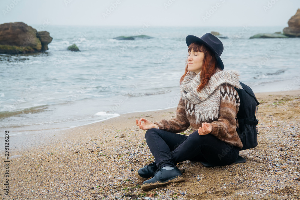 Beautiful red-haired woman in a hat and scarf with a backpack sits in a meditative position on the coast against the background of the rocks against the beautiful sea. Tourism, rest, lifestyle.