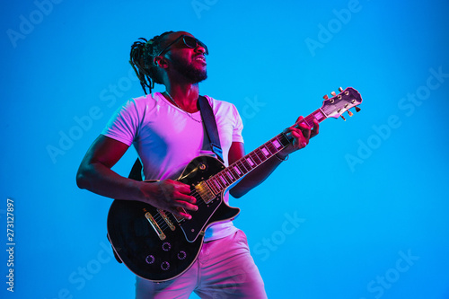 Young african-american musician playing the guitar like a rockstar on blue studio background in neon light. Concept of music, hobby. Joyful attractive guy improvising. Retro colorful portrait. photo