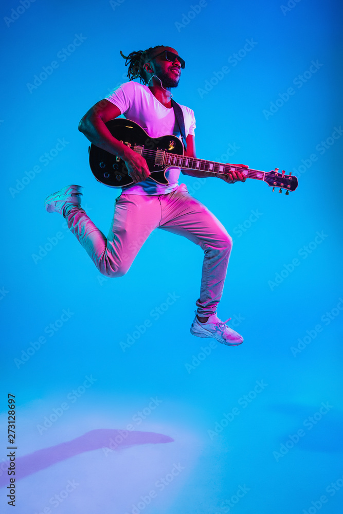Young african-american musician playing the guitar in jump like a rockstar on blue studio background in neon light. Concept of music, hobby. Joyful attractive guy improvising. Retro colorful portrait. Young african-american musician playing the guitar in jump like a rockstar on blue studio background in neon light. Concept of music, hobby. Joyful attractive guy improvising. Retro colorful portrait.
