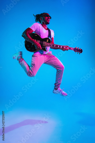Young african-american musician playing the guitar in jump like a rockstar on blue studio background in neon light. Concept of music, hobby. Joyful attractive guy improvising. Retro colorful portrait.