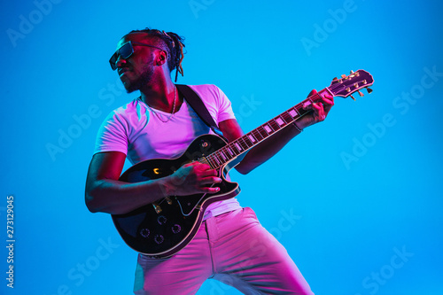 Photography Young african-american musician playing the guitar like a rockstar on blue studio background in neon light
