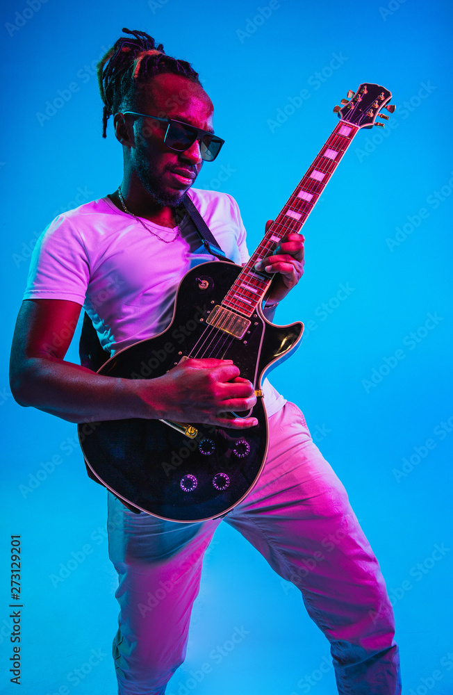 Fototapeta premium Young african-american musician playing the guitar like a rockstar on blue studio background in neon light. Concept of music, hobby. Joyful attractive guy improvising. Retro colorful portrait.