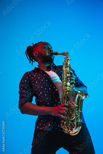 Young african-american jazz musician playing the saxophone on blue studio background in trendy neon light. Concept of music, hobby. Joyful attractive guy improvising. Retro colorful portrait of artist