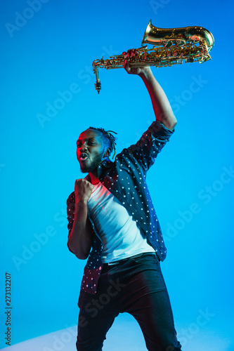 Photography Young african-american jazz musician playing the saxophone on blue studio background in trendy neon light