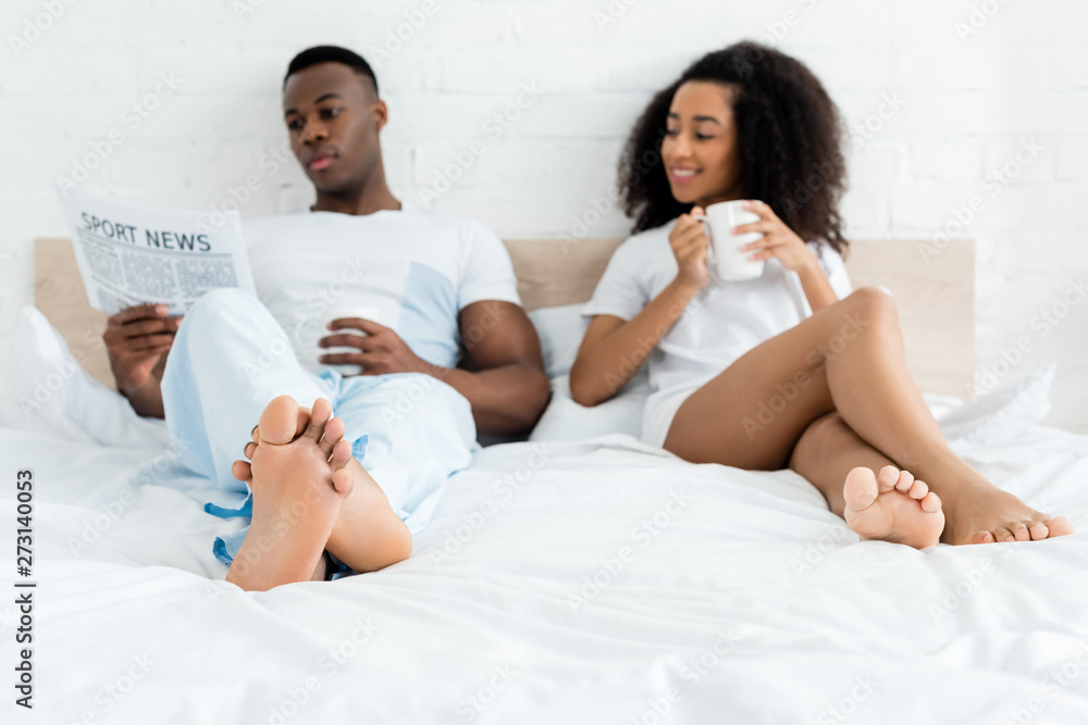 african american woman lying on bed near man with cup and newspaper african american woman lying on bed near man with cup and newspaper