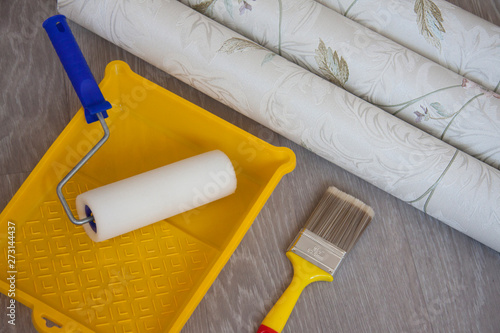 Composition tools for home repair and interior renovation indoors. Rolls of wallpaper, roller and glue container on wooden surface