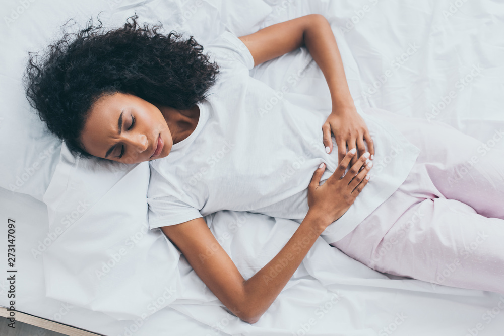 beautiful african american woman suffering from abdominal pain while lying on white bedding beautiful african american woman suffering from abdominal pain while lying on white bedding