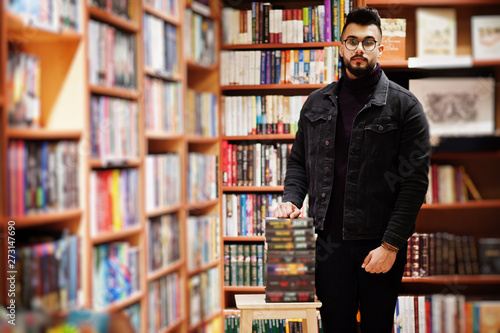 Tall smart arab student man, wear on black jeans jacket and eyeglasses, at library with stack of books.