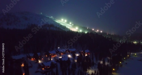 Aerial, reverse, drone shot, away from lighted slopes of Salla fell mountain, overlookiing cabins, in a snowy forest, on polar night, on a dark and moody winter day, in Lapland, Finland photo