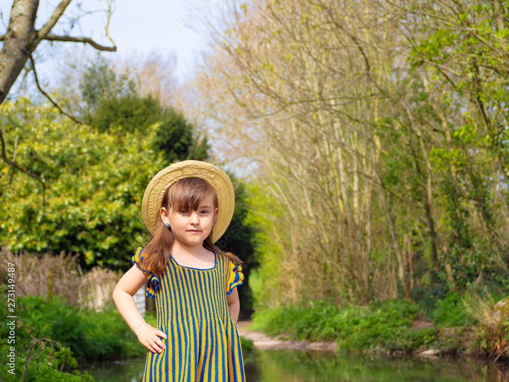 Fototapeta premium Adorable, smiling little girl in rustic dress and straw hat is playing in spring forest