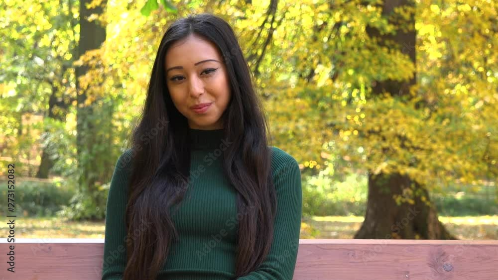 A young Asian woman sits on a bench in a park and smiles at the camera A young Asian woman sits on a bench in a park and smiles at the camera