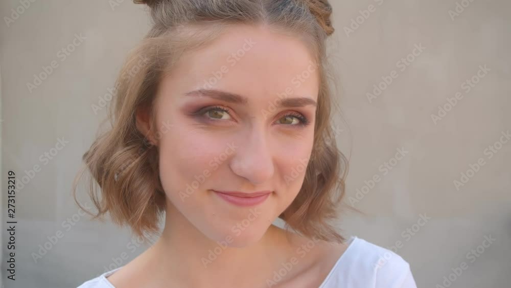 Closeup shoot of young cute caucasian female with hair buns smiling cheerfully flirting looking at camera with urban city on the background Closeup shoot of young cute caucasian female with hair buns smiling cheerfully flirting looking at camera with urban city on the background