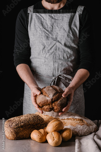 A man in an apron shows an assortment of fresh pastries. Black background.