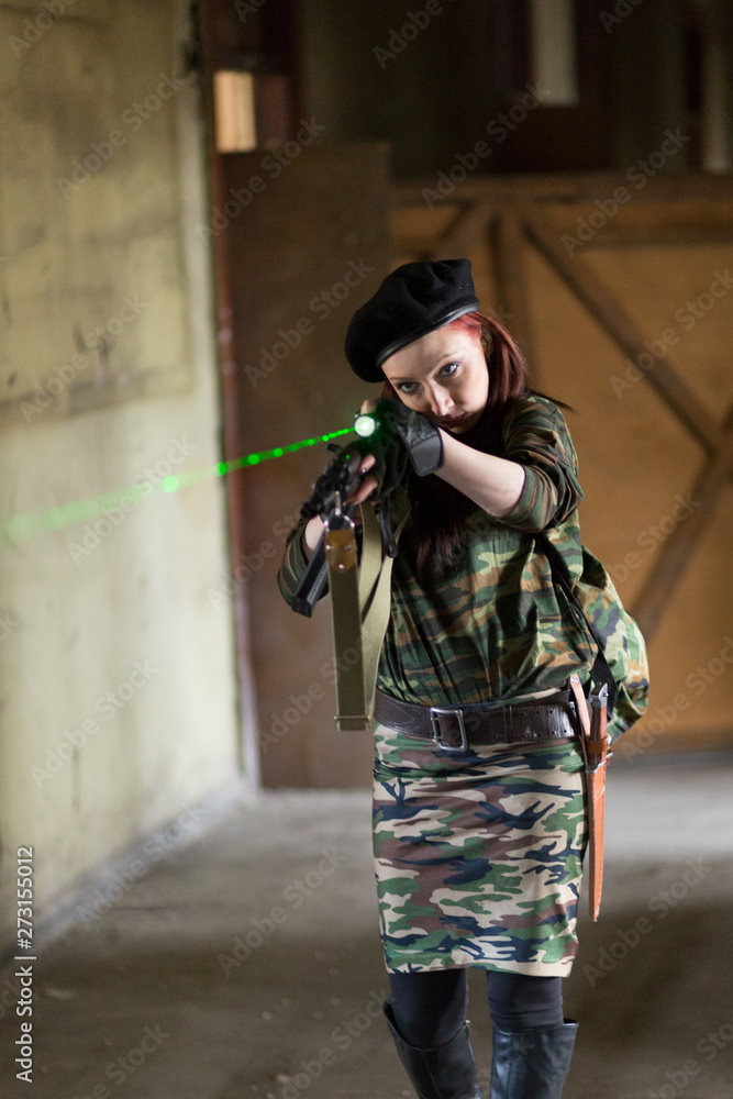 Fototapeta premium A young woman in an abandoned building with a gun. Laser sight on the machine. Aiming from the machine with a laser. The girl in the black beret, in green camouflage.