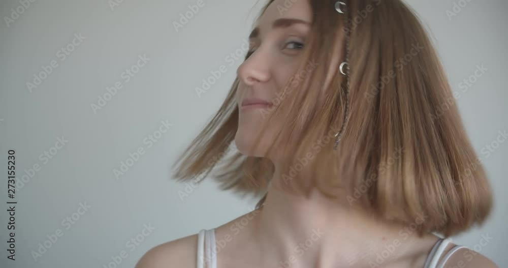 Closeup shoot of young pretty caucasian female with hair rings and glitter makeup smiling happily and tossing her hair turning to camera in the white room indoors Closeup shoot of young pretty caucasian female with hair rings and glitter makeup smiling happily and tossing her hair turning to camera in the white room indoors