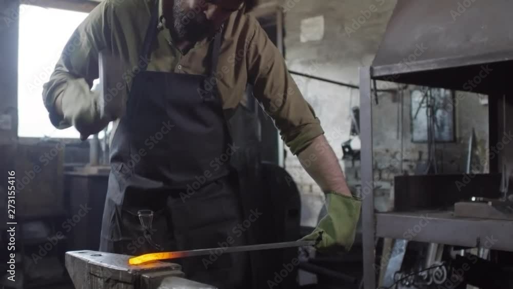Caucasian bearded blacksmith in safety uniform holding candent metal billet in one hand and forging it with hammer with other hand Caucasian bearded blacksmith in safety uniform holding candent metal billet in one hand and forging it with hammer with other hand
