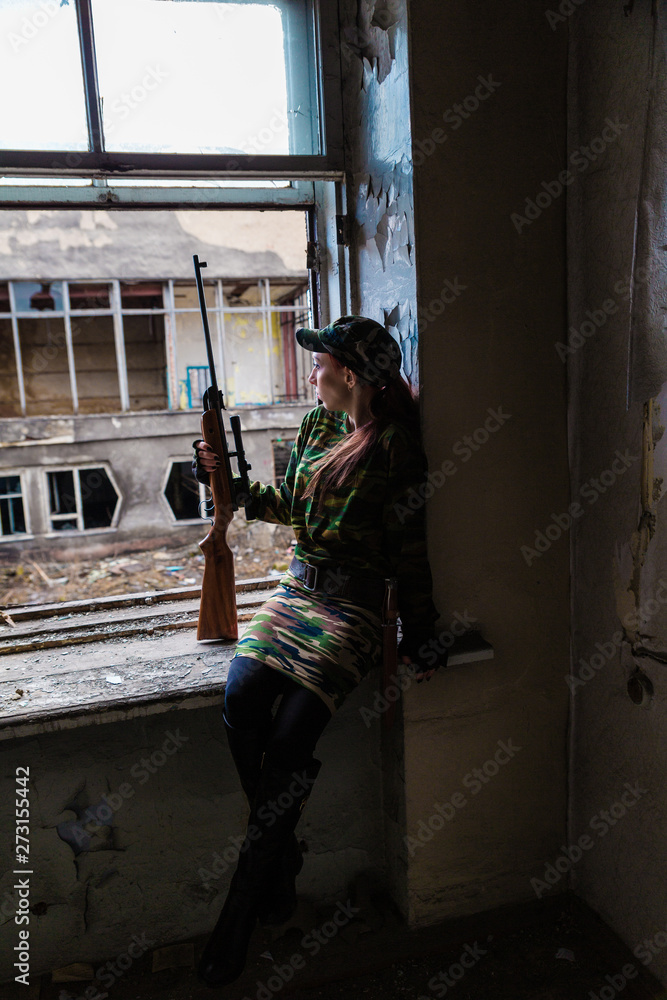 Fototapeta premium A young woman with a rifle in uniform at the window looking at the street. The woman sniper in a green suit and cap.