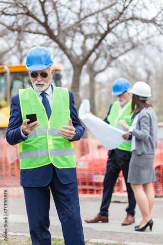 Serious senior engineer or businessman using his smart phone while inspecting a construction site. Two people looking at blueprints, blurred in the background