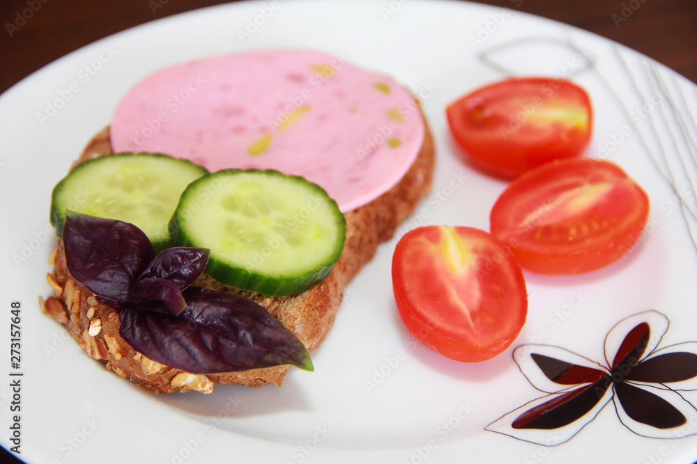 Sandwich of rye bread with delicious sausage, slices of fresh cucumber, tomato and basil leaves on a white plate Sandwich of rye bread with delicious sausage, slices of fresh cucumber, tomato and basil leaves on a white plate