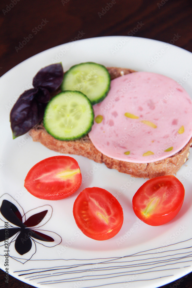 Sandwich of rye bread with delicious sausage, slices of fresh cucumber, tomato and basil leaves on a white plate Sandwich of rye bread with delicious sausage, slices of fresh cucumber, tomato and basil leaves on a white plate