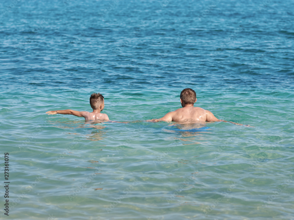 Boy is splashing in the sea with his dad. They are having fun during their holidays on the seacoast. Back view.